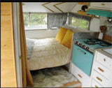 Cozy vintage RV rental interior with a comfy bed, sunny yellow pillows, turquoise stove, and a soft furry rug.
