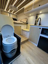 Inside a modern campervan RV rental: kitchenette, raised bed, and portable toilet. Features wood floors & LED lighting.