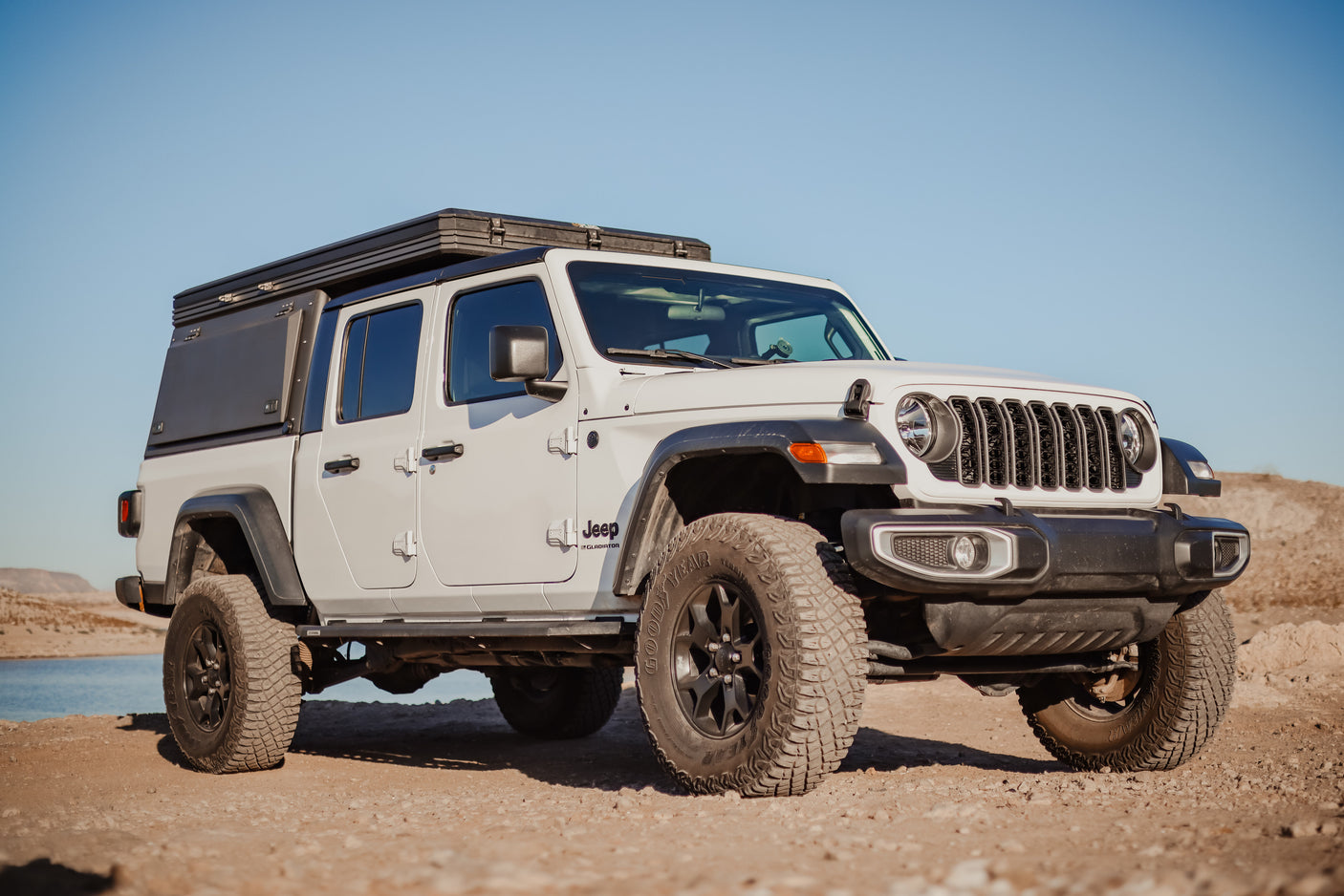 White Jeep Gladiator overland truck with camper on dirt by lake. Ideal for RV rental adventures!