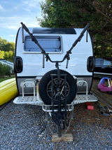 Rear view of a white travel trailer with a bike rack & spare tire, ready for an RV rental adventure. Kayak & child's toy nearby.