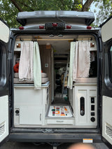 This RV rental shows the functional and cozy interior of a camper van with rear doors open, featuring a bed, kitchen, and controls.
