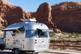 Shiny Airstream rv rental parked in a sunlit desert. Its silver body contrasts with massive red rock formations and a clear blue sky.