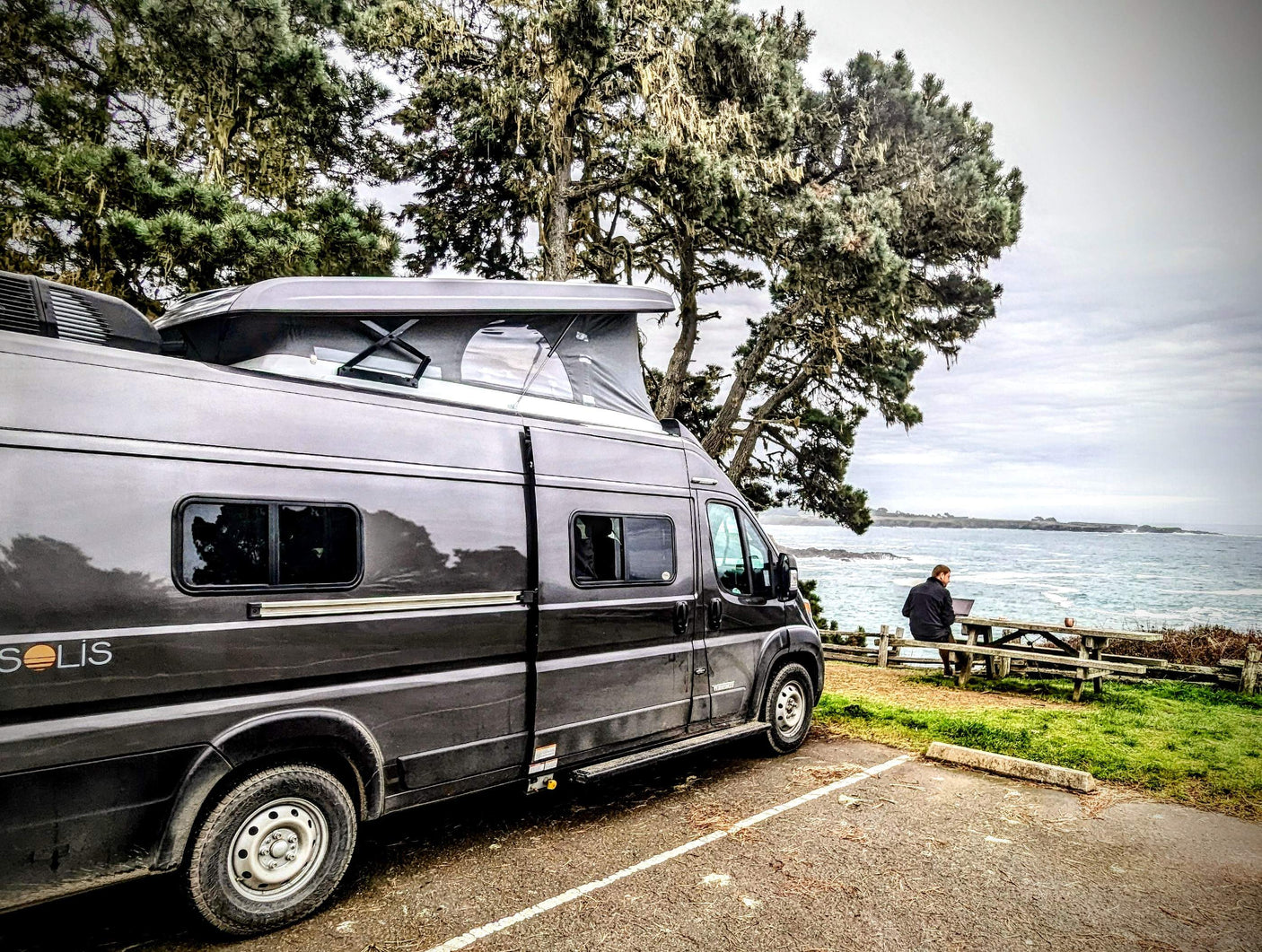 This scenic **rv rental** offers remote work by the ocean. A camper van with a pop-top is parked near a person on a laptop at a picnic table.