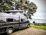 This scenic **rv rental** offers remote work by the ocean. A camper van with a pop-top is parked near a person on a laptop at a picnic table.