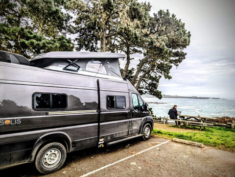 This scenic **rv rental** offers remote work by the ocean. A camper van with a pop-top is parked near a person on a laptop at a picnic table.