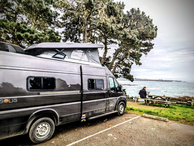 This scenic **rv rental** offers remote work by the ocean. A camper van with a pop-top is parked near a person on a laptop at a picnic table.