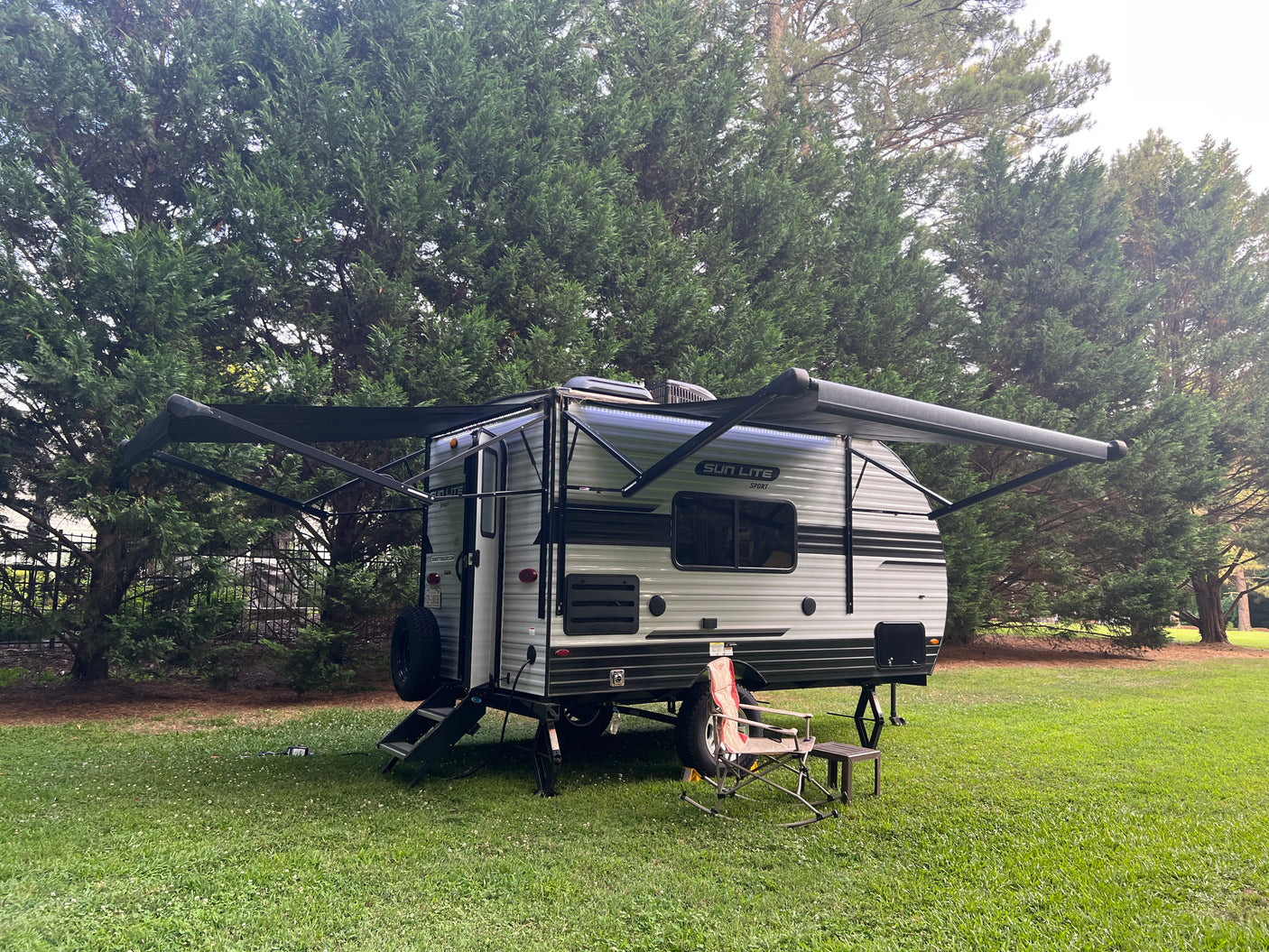 Sun Lite Sport RV rental: A grey travel trailer with extended awnings & LED lights, parked on a green lawn with a chair.