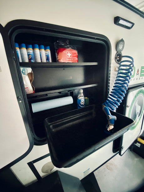 This RV rental's outdoor utility hatch features a sink, faucet, and shelves stocked with sunscreen, a first aid kit, and cleaning supplies.