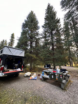 An RV rental camper truck is set up at a scenic forest campsite with a dog, gear, and picnic table.