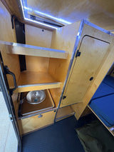 RV rental interior: compact kitchen/storage with wood shelves, sink, LED lights, and blue bed.
