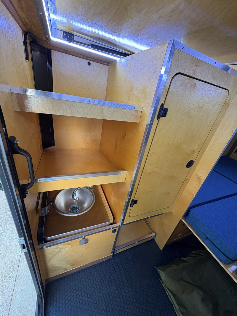RV rental interior: compact kitchen/storage with wood shelves, sink, LED lights, and blue bed. — photo 6