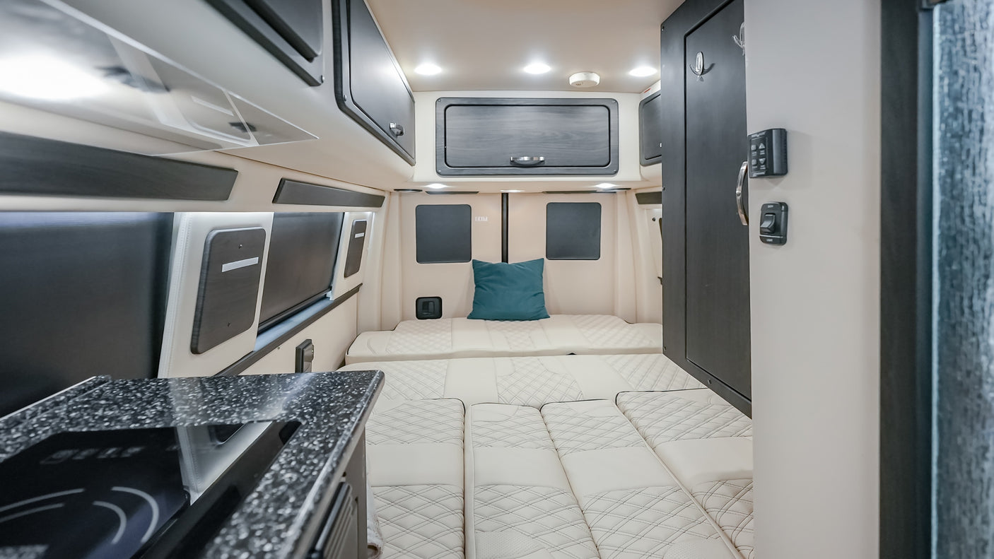 RV rental van interior showcasing modern dark cabinetry, cozy cream quilted bed/seating, and a compact kitchenette.
