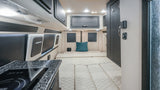 RV rental van interior showcasing modern dark cabinetry, cozy cream quilted bed/seating, and a compact kitchenette.