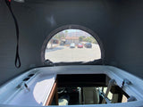 Experience an RV rental! Inside a camper van's pop-top, a bed area with an oval window overlooks a lot, building & mountains.
