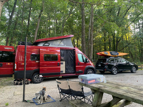 Enjoying an RV rental adventure: Red camper van, kayak, dog, & picnic setup at a beautiful wooded campsite.