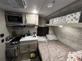 Compact rv rental interior featuring kitchen with microwave, stove, sink, and a comfortable bed with patterned bedding and storage.