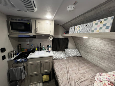 Compact rv rental interior featuring kitchen with microwave, stove, sink, and a comfortable bed with patterned bedding and storage.