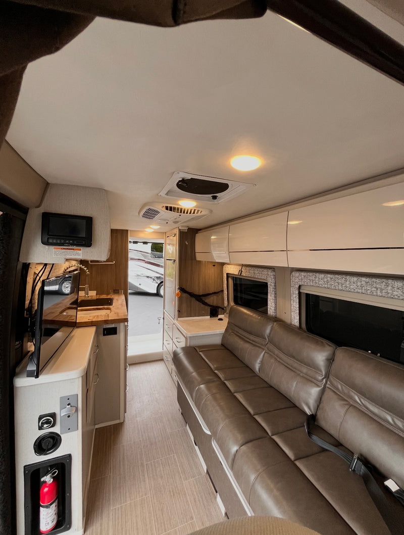 Inside this modern RV rental: a compact kitchen, long brown sofa with seatbelts, overhead cabinets, and bright lighting. — photo 5