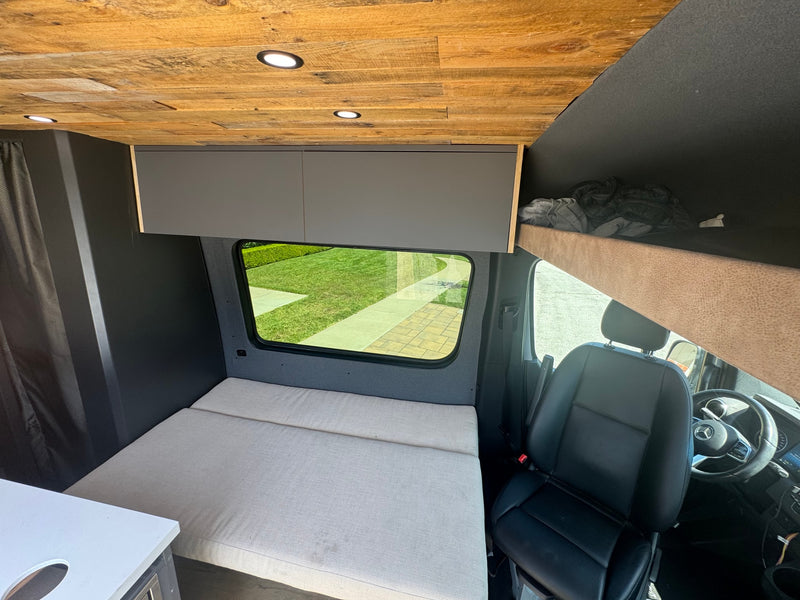 Compact rv rental interior featuring a wooden ceiling, cozy bed, overhead cabinets, and driver's area, ideal for travel. — photo 15