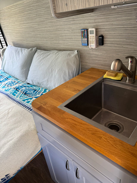 This RV rental features a functional kitchenette with a stainless steel sink, wood counter, light grey cabinets, and a cozy bed.