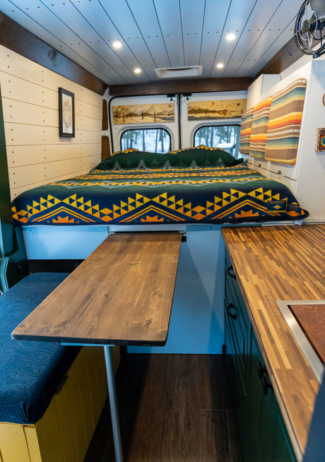 Compact RV rental interior with a comfy bed, pull-out dining table, and built-in kitchen space.