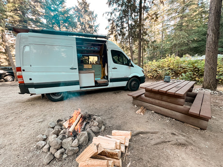 White RV rental van setup at a forest campsite, with a roaring campfire & a wooden picnic table.