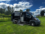 Black camper van RV rental in a vineyard, ready for outdoor living with chairs and table set up on lush grass.