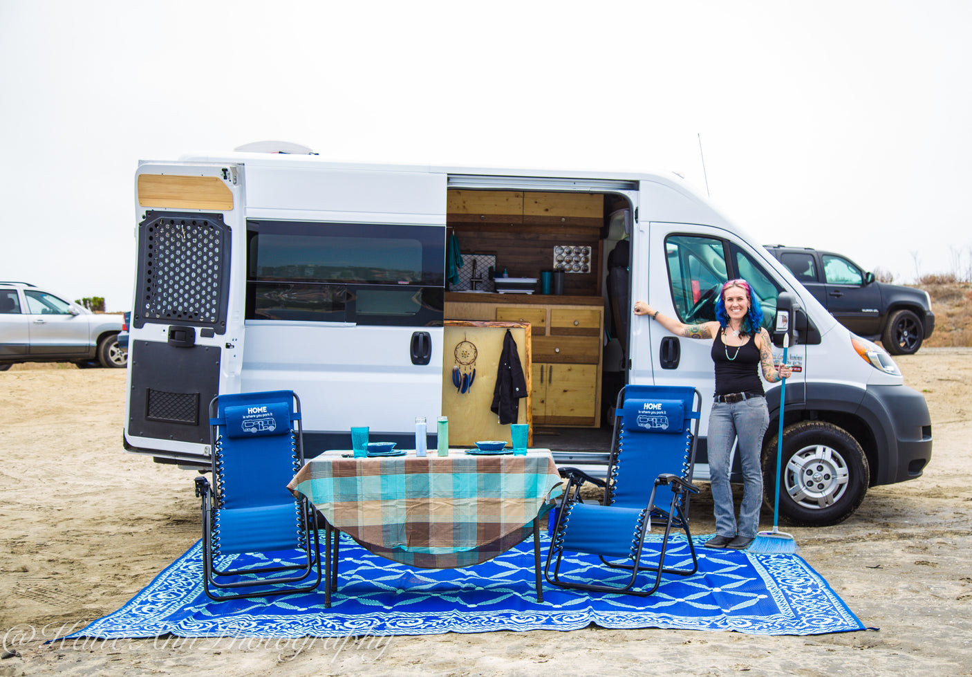 Woman smiles by her RV rental, showcasing the outdoor setup with a table, chairs, and mat on the sand for van life adventures.