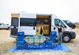 Woman smiles by her RV rental, showcasing the outdoor setup with a table, chairs, and mat on the sand for van life adventures.