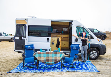 Woman smiles by her RV rental, showcasing the outdoor setup with a table, chairs, and mat on the sand for van life adventures.