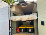 This RV rental camper van interior features a comfy bed, overhead storage, Greenmade storage bins, and a portable shower.