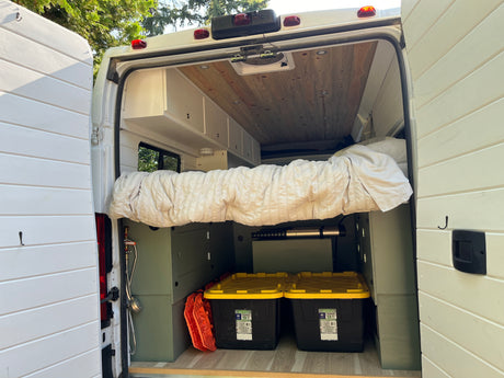 This RV rental camper van interior features a comfy bed, overhead storage, Greenmade storage bins, and a portable shower.