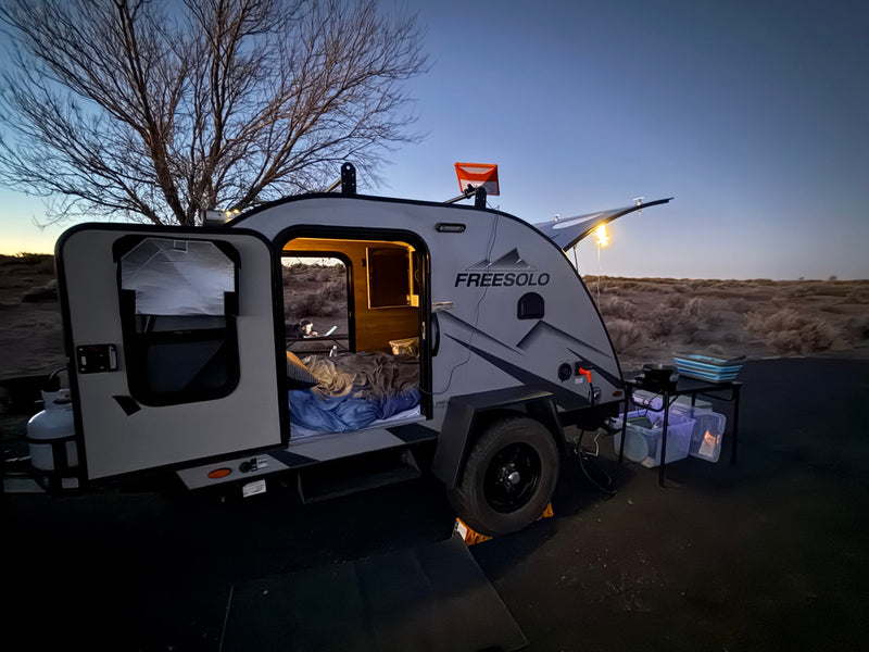 Cozy RV rental camp at dusk: FreeSolo teardrop trailer, open door revealing bed & laptop user, Starlink on top, outdoor cooking. — photo 2