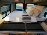Cozy bed in an RV rental with white bedding, patterned blanket, and comfy pillows, set against scenic rear windows.