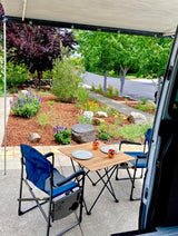 This RV rental features an outdoor patio with a wooden table, chairs, and a landscaped garden. Ideal for leisure.