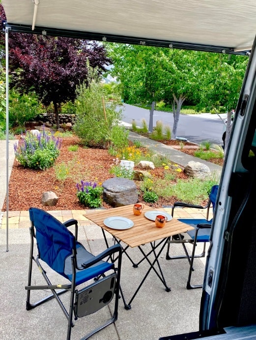 This RV rental features an outdoor patio with a wooden table, chairs, and a landscaped garden. Ideal for leisure. — photo 17