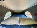 This RV rental's cozy pop-top sleeping area features a bed with pillows and side windows offering views of trees.