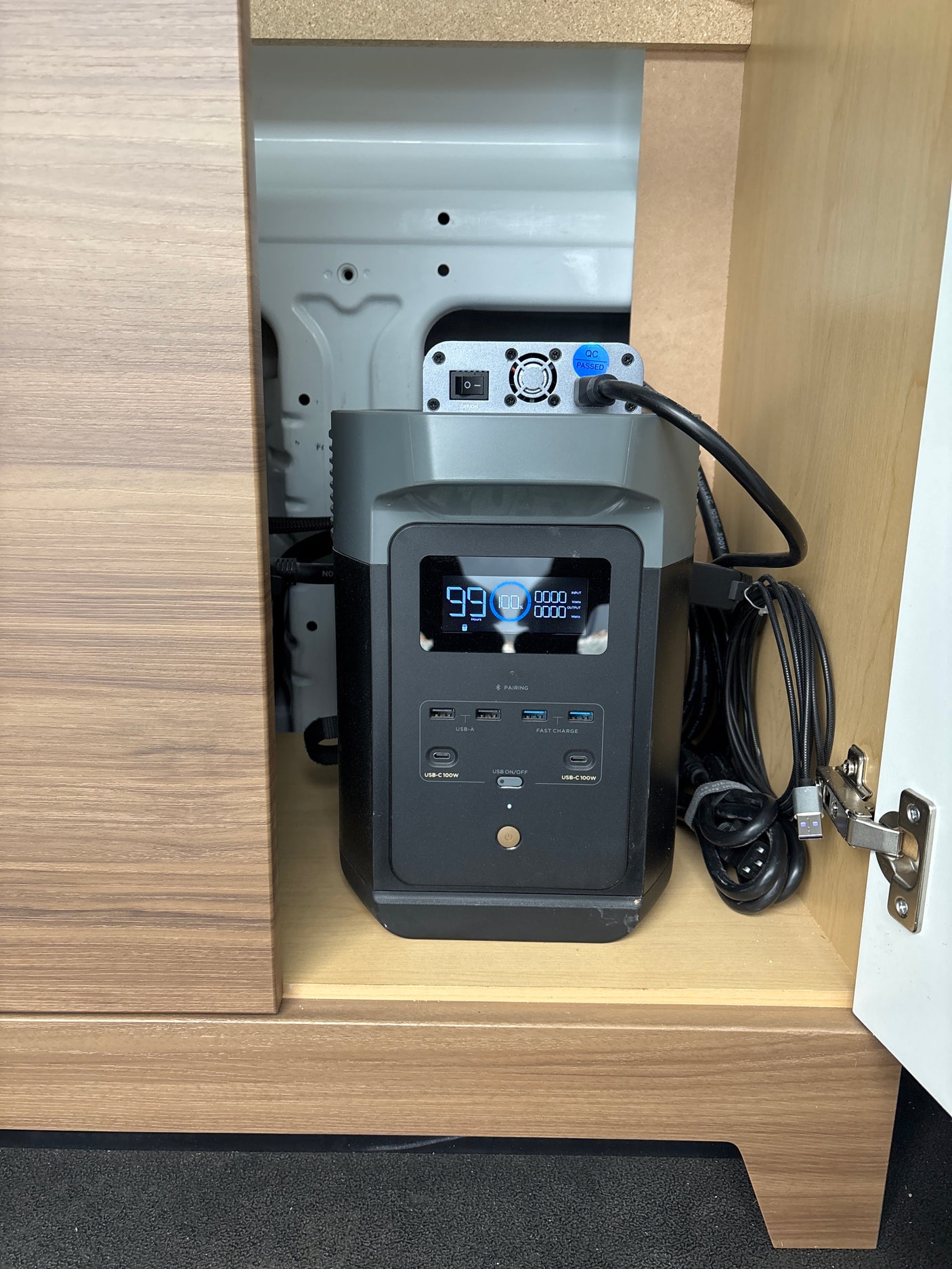 A portable power station showing 99% charge, with a top-mounted solar controller, neatly installed inside a wooden cabinet for an RV rental.