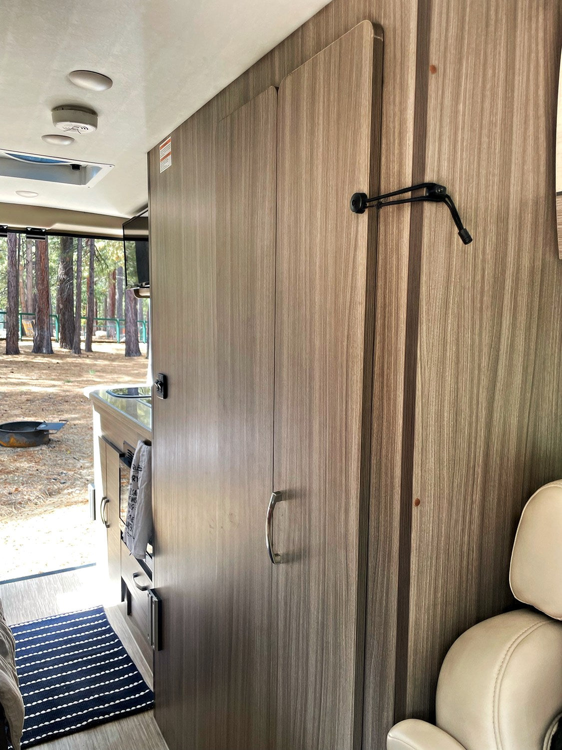 Inside this RV rental, light wood-grained cabinets lead to a kitchenette with a window view of pine trees at a campsite.