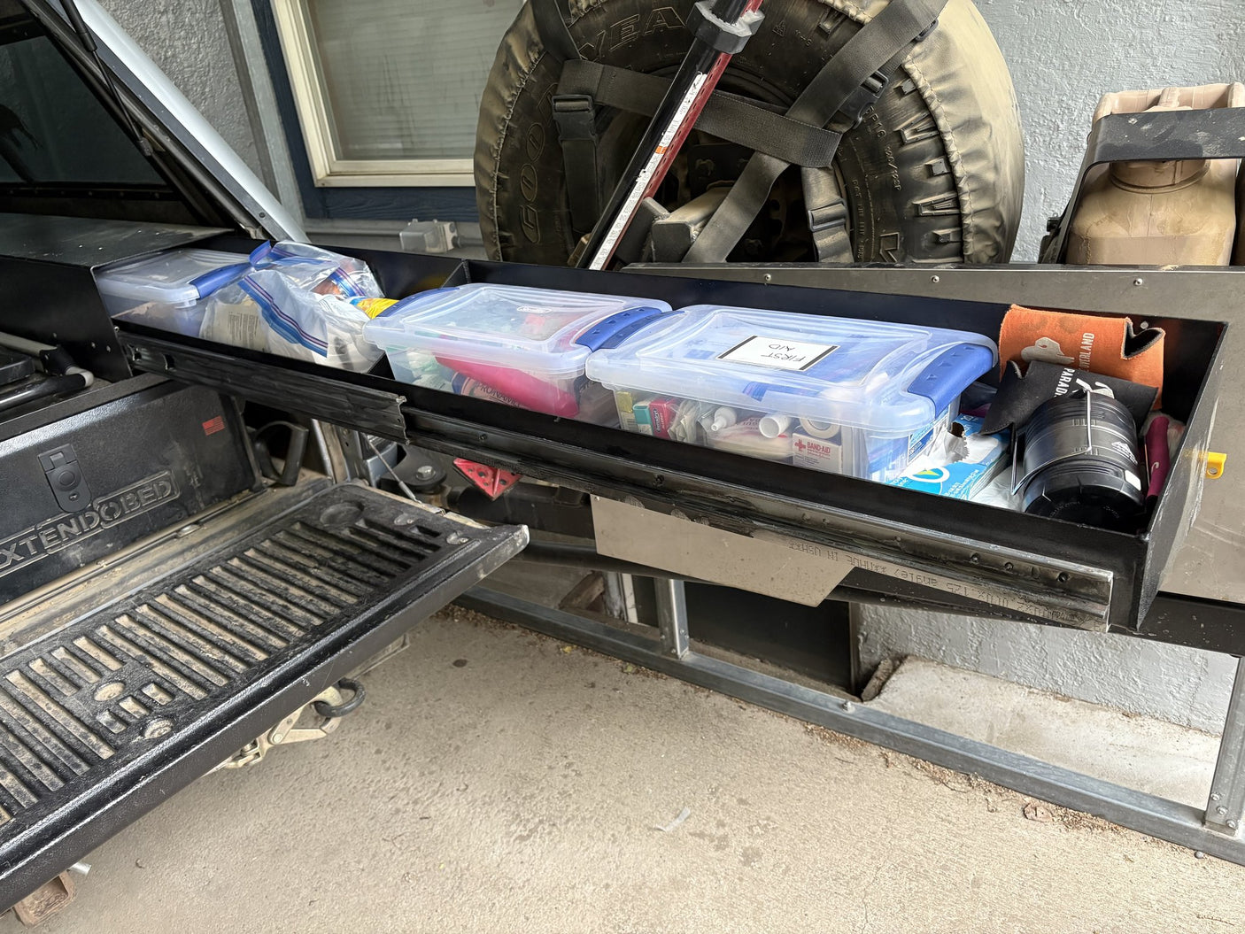 Long side drawer of the Extendobed system pulled open, loaded with clear storage bins including a first aid kit, next to the open tailgate