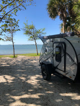 Waterfront camping with an RV rental! A grey FreeSolo teardrop camper is parked on a shaded path by the bay, surrounded by trees.