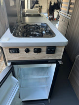 An rv rental's compact kitchen features a two-burner stove, sink with faucet, and an open mini-fridge with shelves.