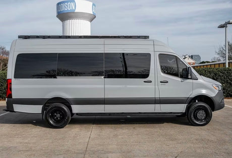 Side view of a light gray Mercedes-Benz Sprinter camper van, perfect for rv rental. Customized with a roof rack and black wheels.