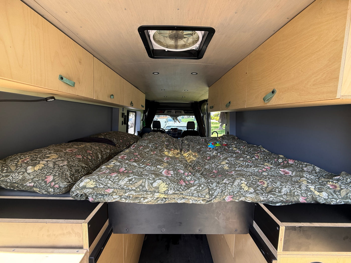 Cozy RV rental camper van interior featuring a large bed with floral bedding, wooden overhead cabinets, and a ceiling fan. — photo 8