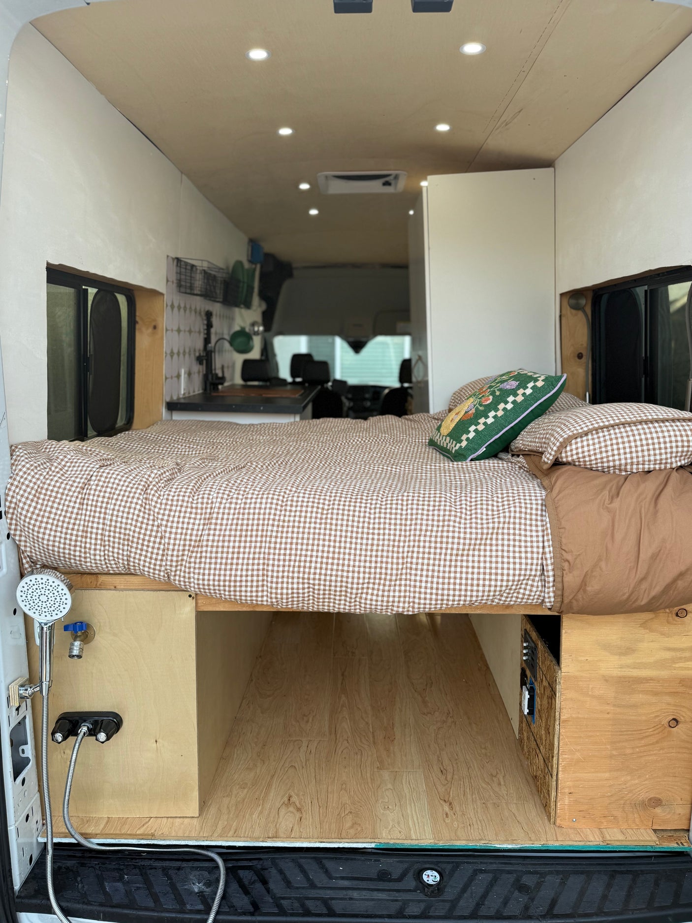 Inside a modern rv rental camper van: comfortable bed with gingham bedding, kitchen, and outdoor shower setup.