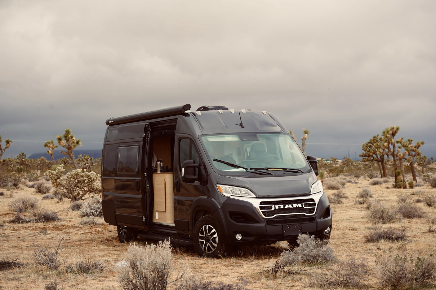 Dark grey Ram Promaster RV rental camper van parked in a desert with Joshua trees under an overcast sky, side door open.