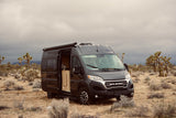 Dark grey Ram Promaster RV rental camper van parked in a desert with Joshua trees under an overcast sky, side door open.