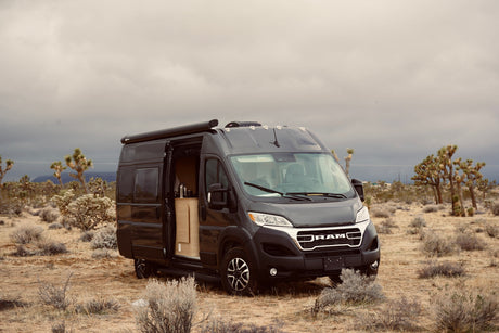 Dark grey Ram Promaster RV rental camper van parked in a desert with Joshua trees under an overcast sky, side door open.