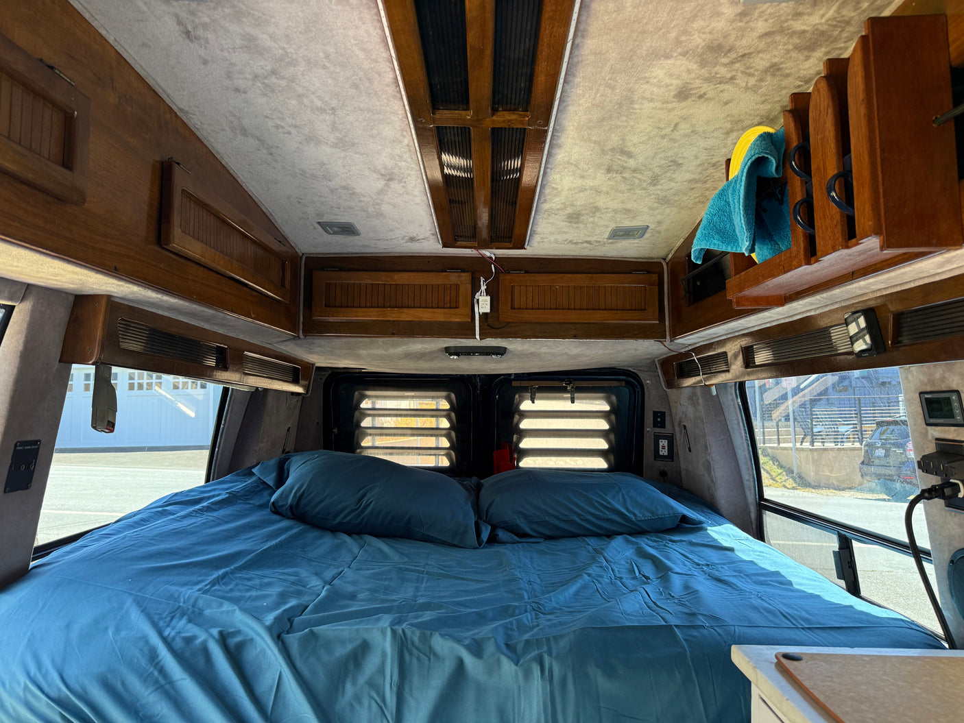 Interior of an RV rental camper van, showing a neatly made bed with blue sheets, wooden overhead cabinets, and rear window blinds.