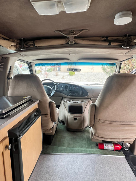 Inside this rv rental, view forward: tan interior, driver & passenger seats, dashboard, and a compact kitchen setup on left.